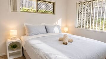 A neatly made bed with a rolled towel, two bedside lamps, a small plant on a nightstand, and a window with a metal screen.