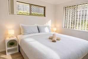 Ein ordentlich bezogenes Bett mit einem aufgerollten Handtuch, zwei Nachttischlampen, einer kleinen Pflanze auf einem Nachttisch und einem Fenster mit einem Metallfliegengitter.