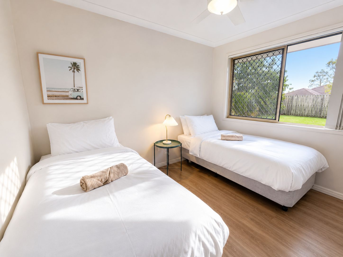 A bedroom with two single beds, a nightstand with a lamp, a framed picture on the wall, and a window with a view of the outdoors.
