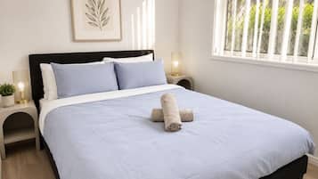 Een net opgemaakt bed met een blauwe sprei, twee kussens en een opgerolde handdoek. Aan de muur hangt een ingelijst botanisch kunstwerk, bij het bed staat een nachtkastje met een lamp en er is een raam met uitzicht op groen.