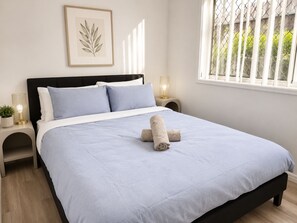 A neatly made bed with a blue bedspread, two pillows, and a rolled towel. There is a framed botanical print on the wall, a bedside table with a lamp, and a window with a view of greenery. - 4 BR Family Retreat – 15 min to Ipswich (Redbank Plains)