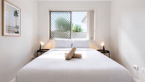 A neatly made bed with a rolled towel, two bedside lamps, a window with a view of greenery, and a framed picture on the wall. - 4 BR Family Retreat – 15 min to Ipswich (Redbank Plains)
