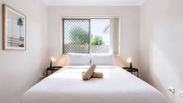 A neatly made bed with a rolled towel, two bedside lamps, a window with a view of greenery, and a framed picture on the wall.