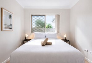 Ein ordentlich bezogenes Bett mit einem zusammengerollten Handtuch, zwei Nachttischlampen, ein Fenster mit Blick ins Grüne und ein gerahmtes Bild an der Wand.