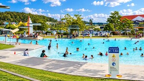Ein öffentliches Schwimmbad unterschiedlicher Wassertiefe, umgeben von einem Parkplatz und einer Rasenfläche.