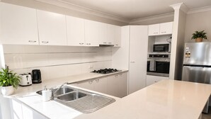 A modern kitchen with white cabinetry, a stainless steel sink, a gas stove, a microwave, and a refrigerator. - 4 BR Family Retreat – 15 min to Ipswich (Redbank Plains)