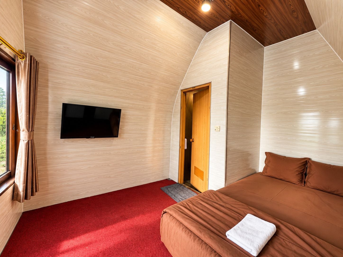 Ein Zimmer mit einem an der Wand montierten Flachbildfernseher, einer Holztür, einem roten Teppich und einem Bett mit braunen Bettwaren.
