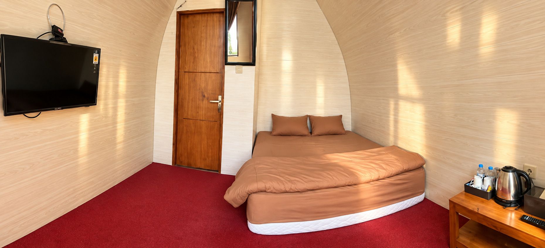 A bedroom with a bed, a television mounted on the wall, a wooden door, and a small table with items on it.
