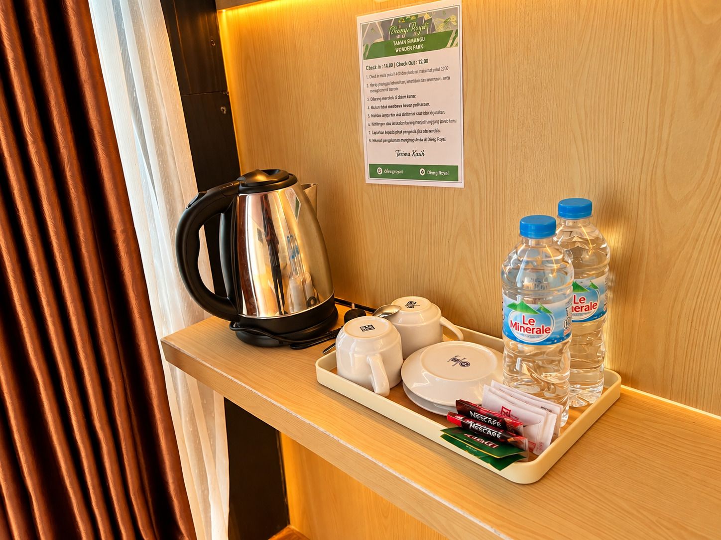 Un plateau contenant une bouilloire en acier inoxydable, des bouteilles d’eau et des sachets de Nescafe, posé sur une surface en bois.