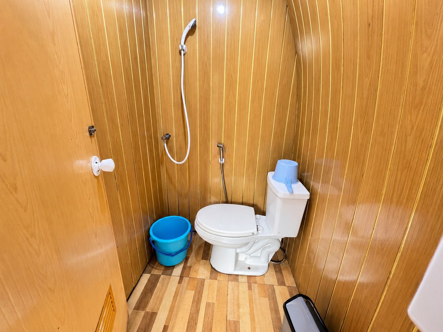 Un baño pequeño con pared de paneles de madera, un inodoro blanco, un cubo azul y una manguera de ducha.