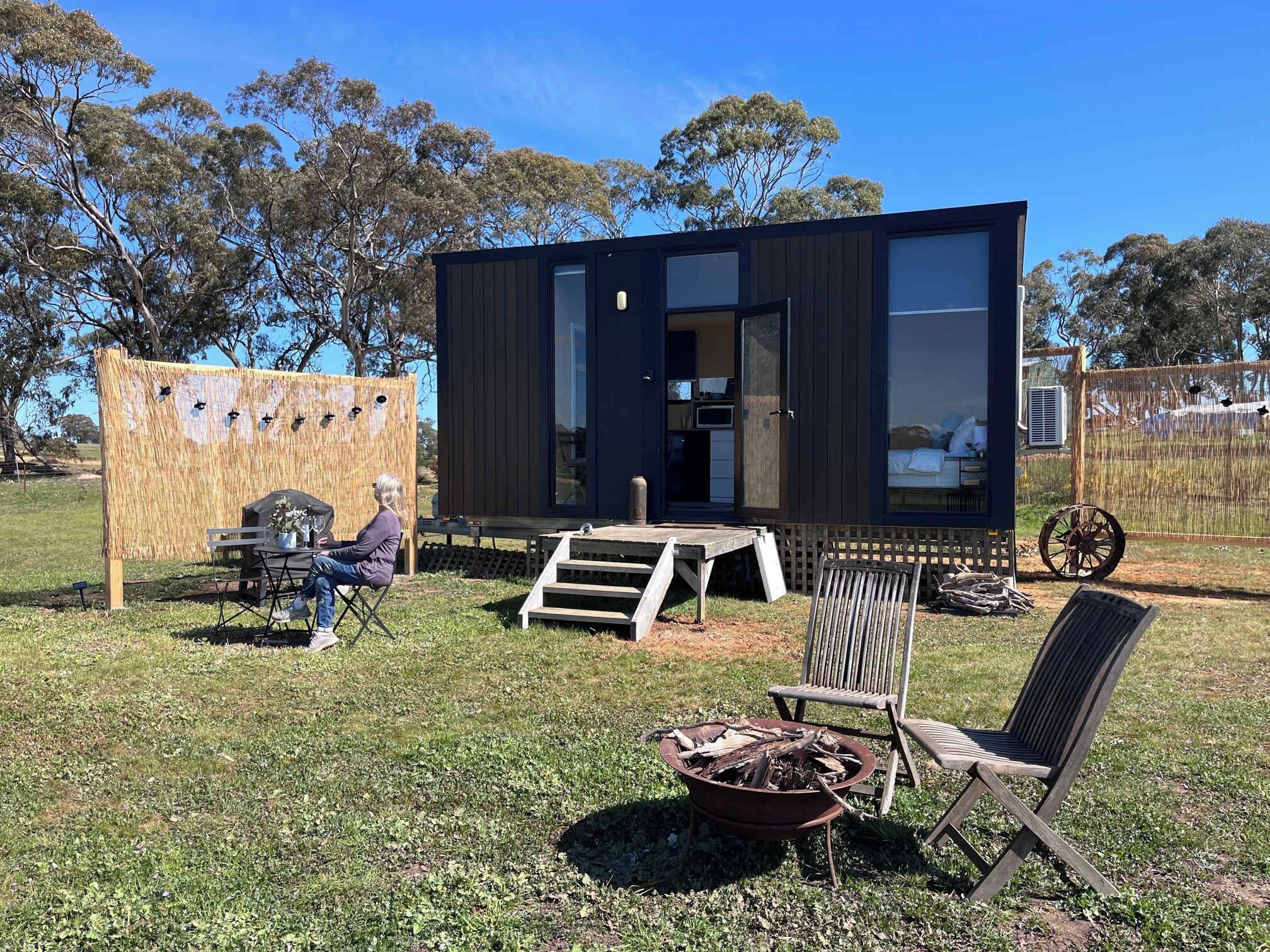 A modern, compact cabin with a large window, a small deck, and a fire pit in the yard.