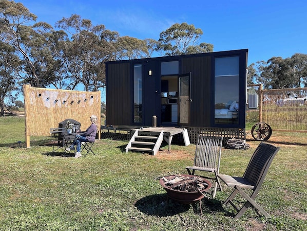 A modern, compact cabin with a large window, a small deck, and a fire pit in the yard. - Stringybark by Tiny Away (Rhymney)