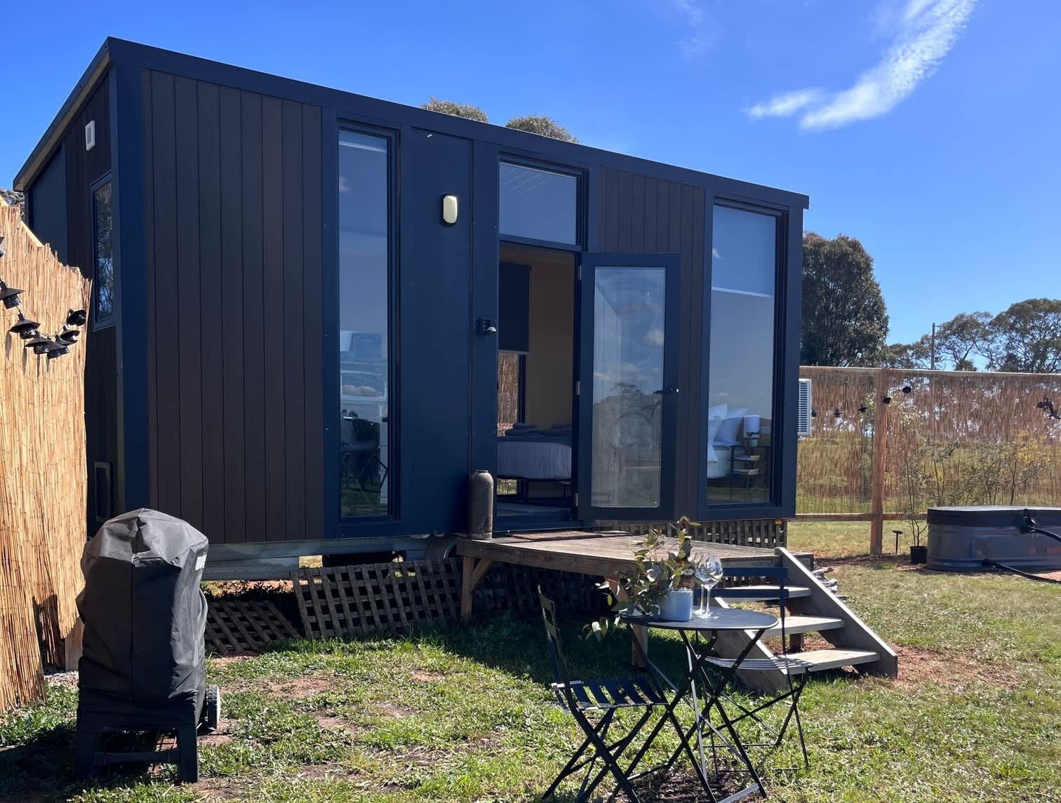 Une maison mobile moderne, de couleur sombre, dotée de grandes portes vitrées, d’une petite terrasse et d’un espace patio avec une table et des chaises.