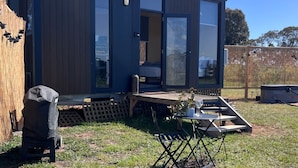 A modern, dark-colored mobile home with large glass doors, a small deck, and a patio area with a table and chairs. - Stringybark by Tiny Away (Rhymney)