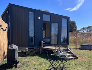 A modern, dark-colored mobile home with large glass doors, a small deck, and a patio area with a table and chairs. - Stringybark by Tiny Away (Rhymney)