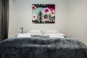 Een net opgemaakt bed met witte kussens en een grijze gebreide deken, een wanddecoratie met bloem- en vlinderdesigns, en een klein nachtkastje met een lamp.