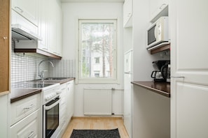 A compact kitchen with white cabinetry, a built-in oven, a microwave, and a window with blinds. - City Station Flat By Lapland Host (Rovaniemi)