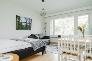 A modern bedroom with a bed, a sofa, a dining table with chairs, and a large window with blinds. - City Station Flat By Lapland Host (Rovaniemi)