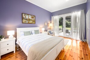 Une chambre à coucher avec un lit, des tables de chevet, une lampe, un tableau au mur et une vue sur l’extérieur, visible à travers une porte vitrée.