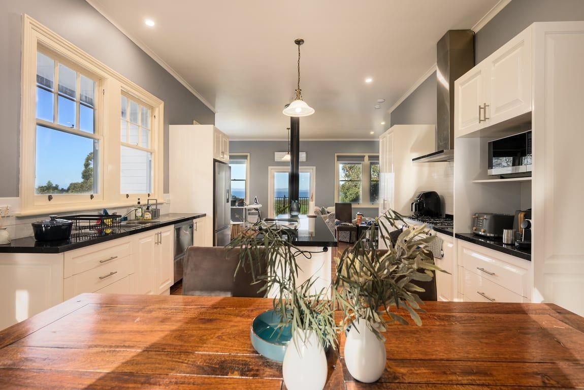 A modern kitchen with white cabinetry, a wooden dining table, and a view of the outdoors through large windows.