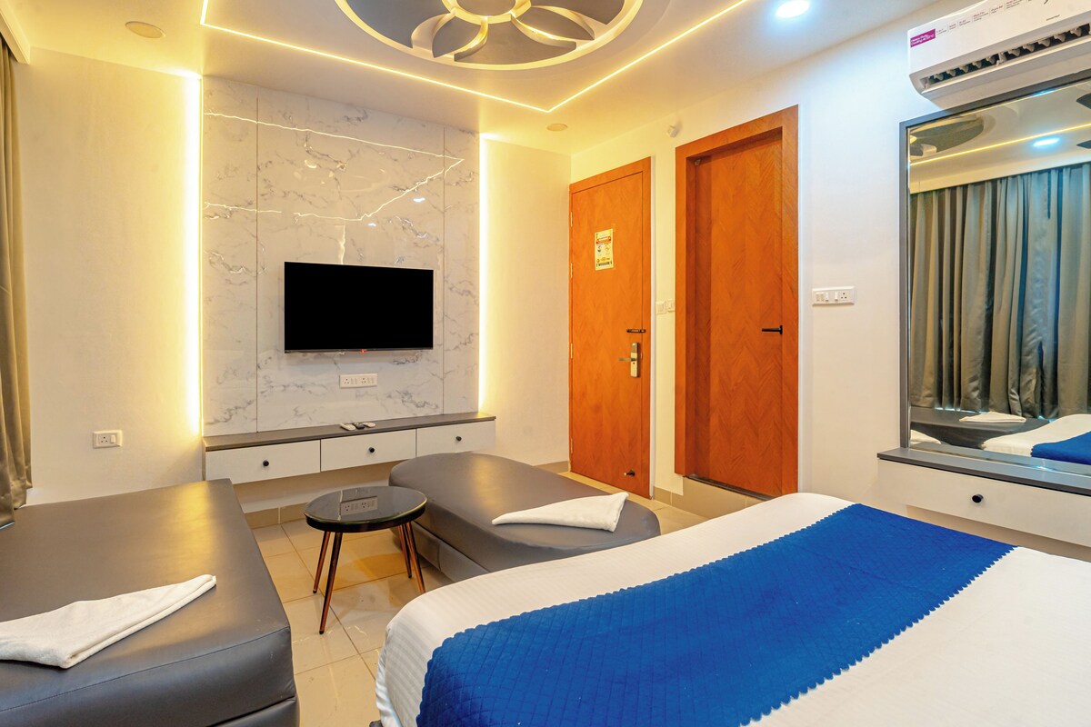 Habitación de hotel con televisor de pantalla plana montado en una pared de mármol, una camilla de masaje, una cama con manta azul y un espejo grande que refleja la habitación.