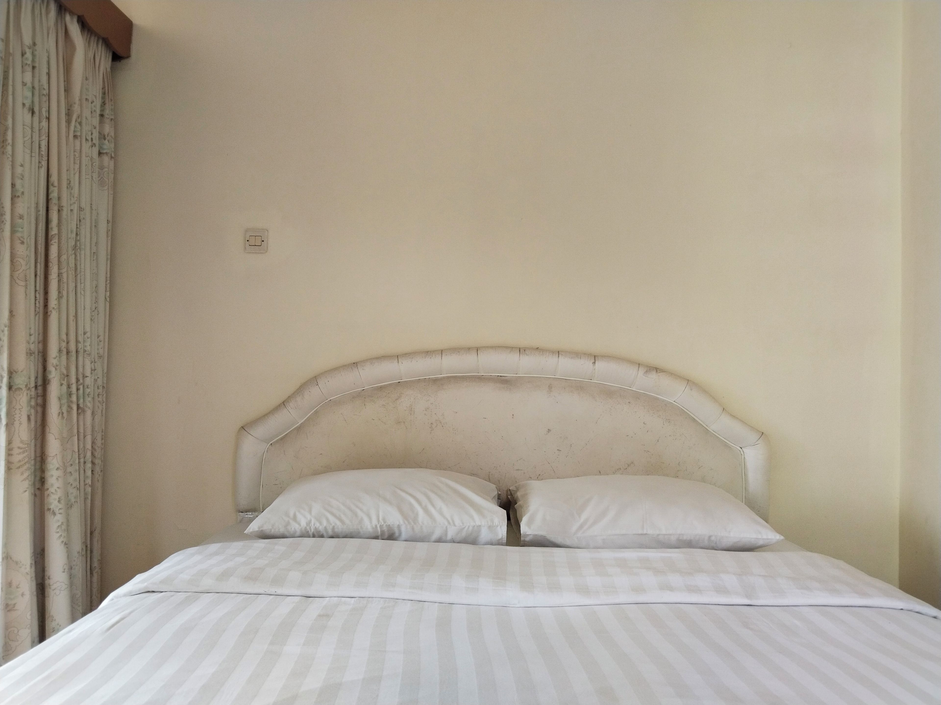 Ein Bett mit Kopfteil, zwei Kissen und einer gestreiften Bettdecke in einem Zimmer mit schlichten Wänden und einem Vorhang.