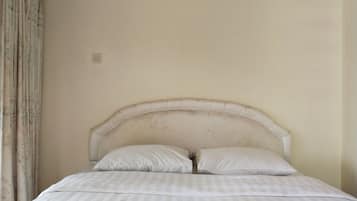 Ein Bett mit Kopfteil, zwei Kissen und einer gestreiften Bettdecke in einem Zimmer mit schlichten Wänden und einem Vorhang.
