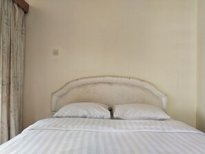 A bed with a headboard, two pillows, and a striped bedspread in a room with plain walls and a curtain.
