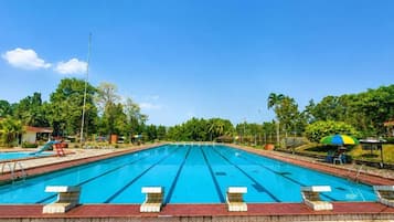 Una amplia piscina al aire libre con varias pistas, rodeada por un solárium de piedra y follaje.