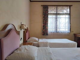 Una habitación con dos camas, una mesita de noche con lámpara, un ventanal con cortinas estampadas y un cabecero de madera.