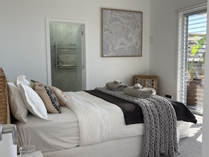 Een net opgemaakt bed met kussens en een dekbed, een handdoekrek, een kunstwerk aan de muur en een raam met jaloezieën.