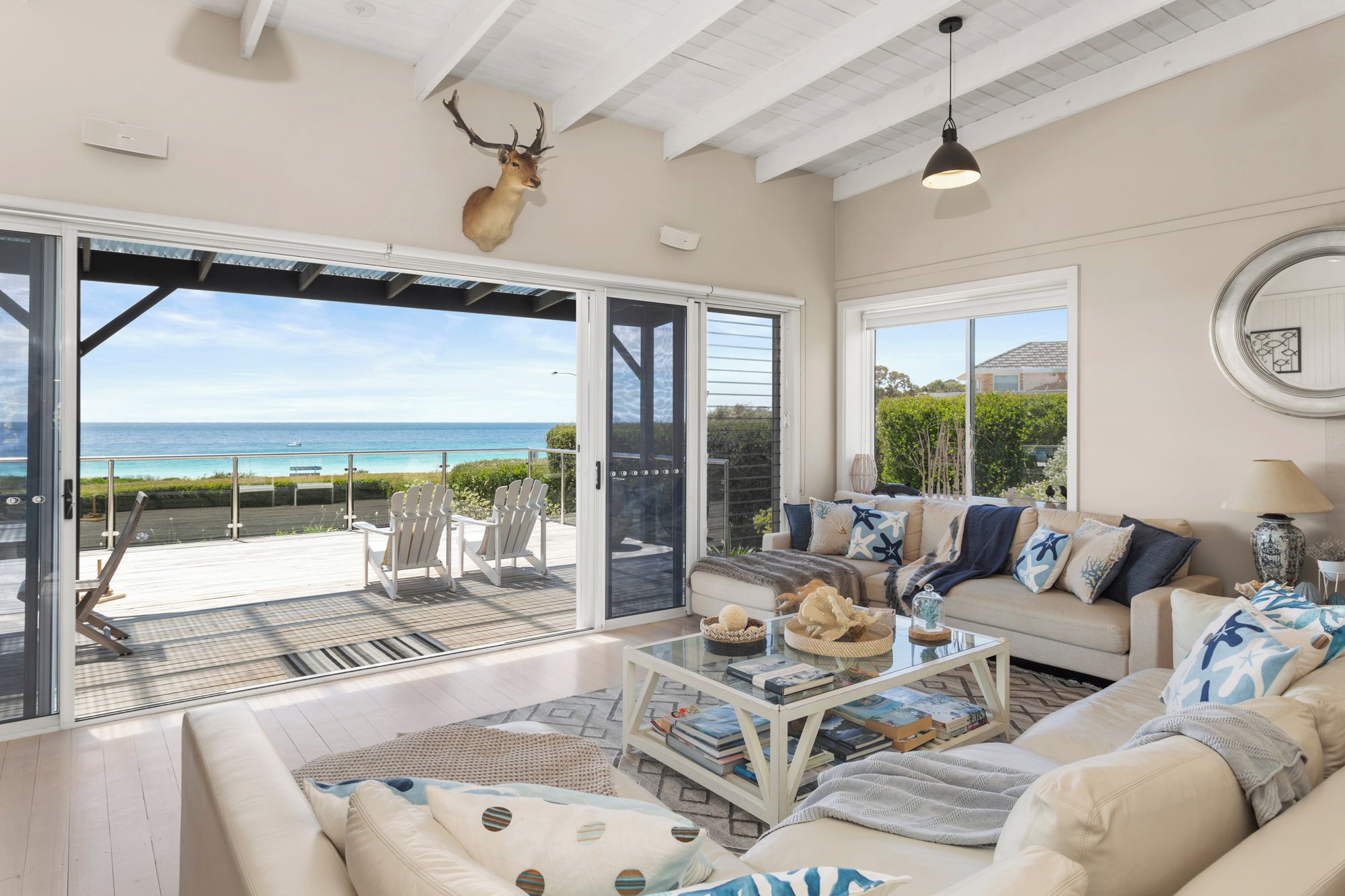 A spacious living room with a large glass door leading to a deck overlooking the ocean. There is a mounted deer head on the wall, a sofa with nautical-themed pillows, and a coffee table with books and decorative items.
