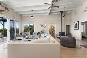 A spacious living room with a large sofa, a coffee table, and a fireplace. There are multiple windows providing natural light and views of the outdoors.
