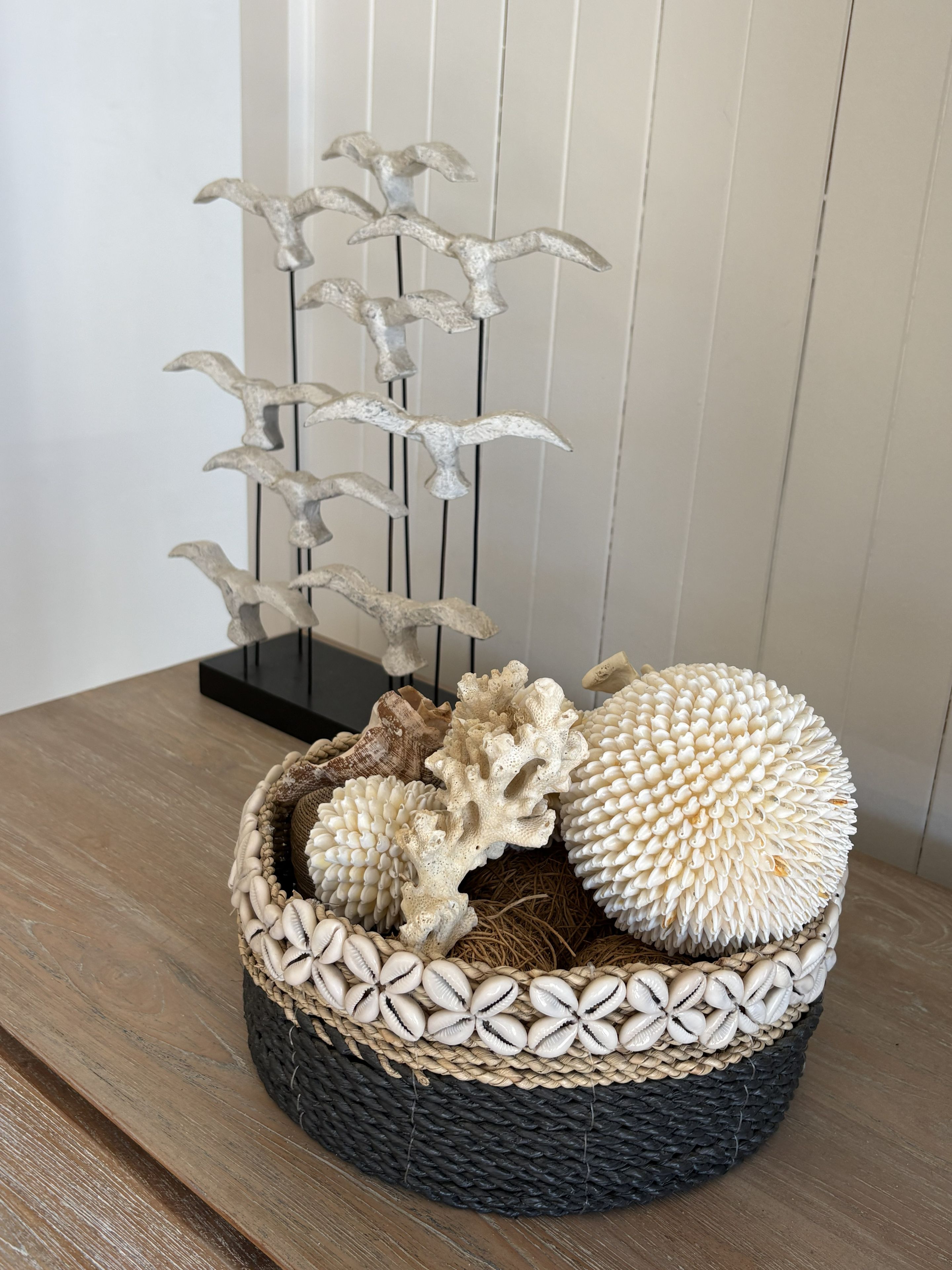 Een gevlochten mand met decoratieve schelpen en een metalen sculptuur op een houten oppervlak.
