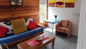A room with a wooden wall, a blue sofa with colorful cushions, a small table with a bowl, a chair, and a painting of birds. - Water Brook 2 B/R Studio Apt (Hamilton)