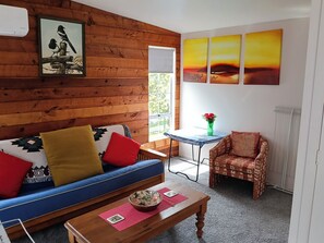 A room with a wooden wall, a blue sofa with colorful cushions, a small table with a bowl, a chair, and a painting of birds. - Water Brook 2 B/R Studio Apt (Hamilton)