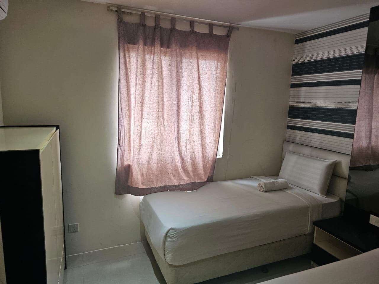 A single bed with a pillow, a bedside table, a window with curtains, and a wall with a striped pattern.