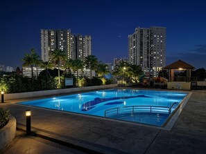 A swimming pool with a surrounding deck, palm trees, and high-rise buildings in the background.