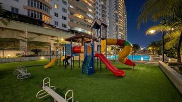 An outdoor playground with slides and exercise equipment, surrounded by a pool and palm trees, with a high-rise building in the background.
