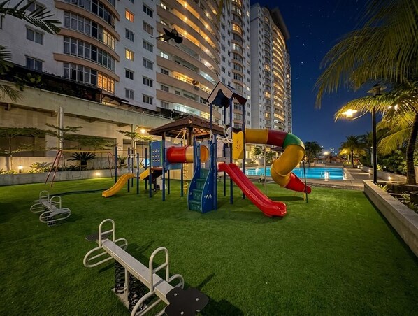 An outdoor playground with slides and exercise equipment, surrounded by a pool and palm trees, with a high-rise building in the background.