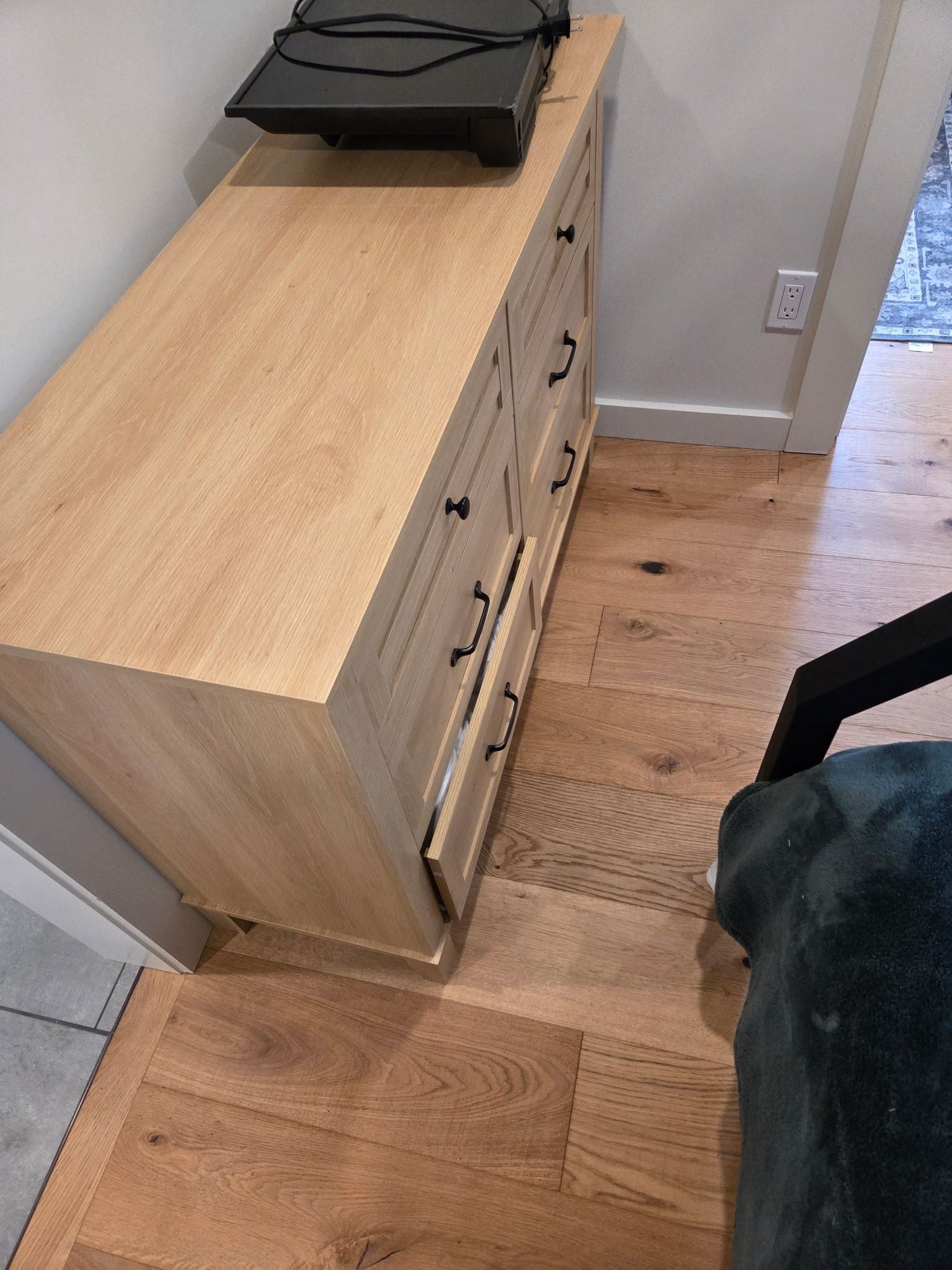 Un comò in legno con cassetti e un piano, una presa elettrica sulla parete e un tappeto decorato sul pavimento.