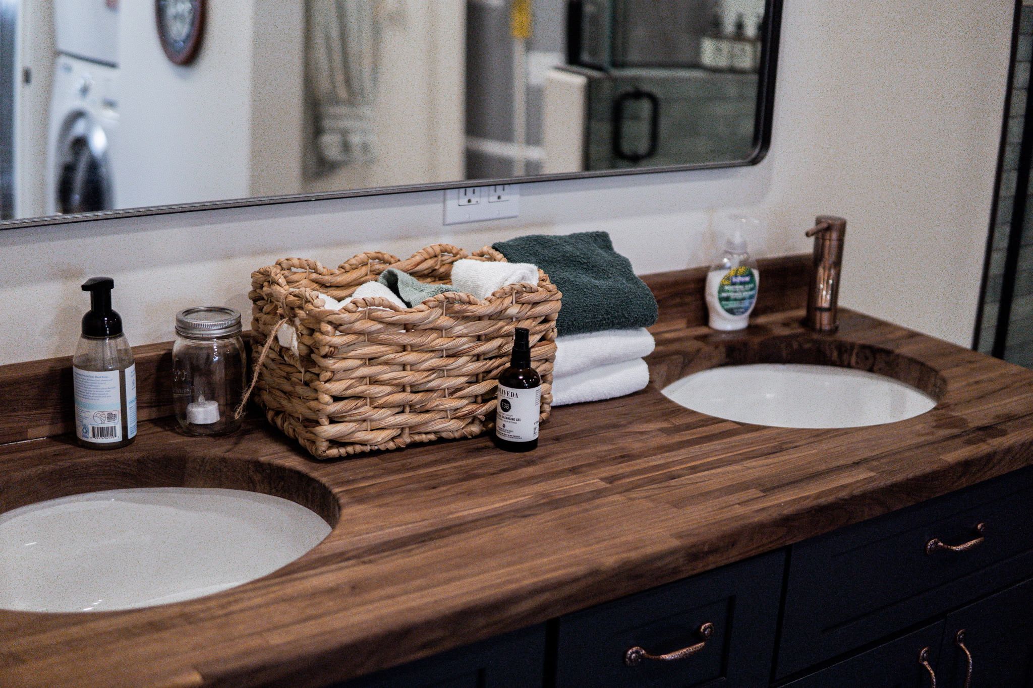 A bathroom vanity with two sinks, a mirror, a wicker basket, a bottle of hand soap, a jar, and a bottle of cleaning solution.