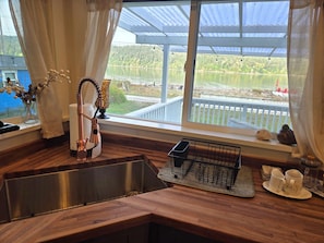 A kitchen with a stainless steel sink, a dish rack, and a view of a lake through a large window.