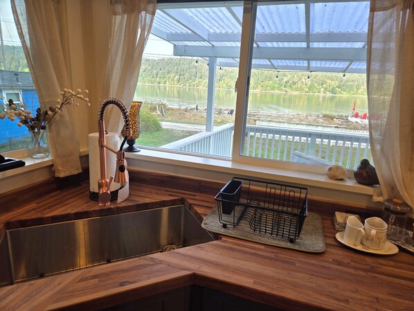A kitchen with a stainless steel sink, a dish rack, and a view of a lake through a large window.