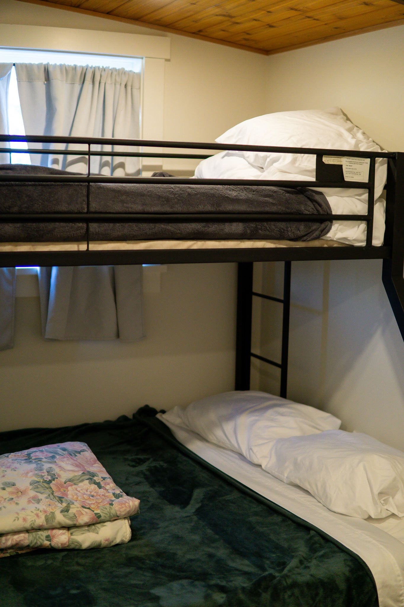 A bunk bed with a window above, a bed with a floral blanket, and a wall with a wooden ceiling.