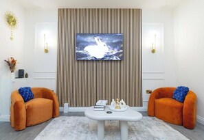 A living room with a flat-screen TV mounted on a wall, two orange armchairs, a white coffee table, and a striped wall panel. - Cosy 3 Bedroom House In Manchester Sleeps 7 (Salford)