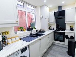 A modern kitchen with white cabinetry, a black stove, and a window with a view of a brick building. - Cosy 3 Bedroom House In Manchester Sleeps 7 (Salford)