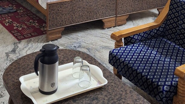 Quarto de hotel com cama, mesa com chaleira e copos, e poltrona estampada.