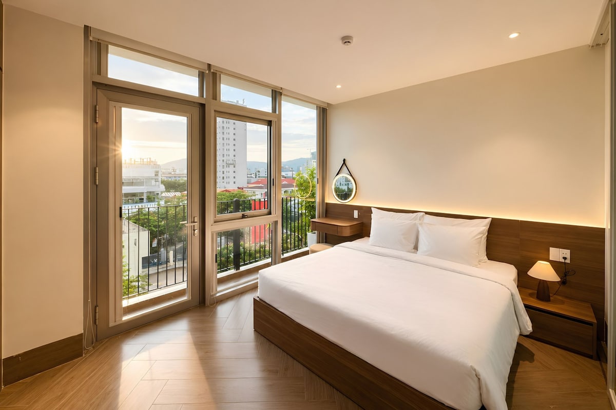 A hotel room with a large bed, a nightstand, a lamp, and a view of the city through large windows.