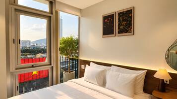 A hotel room with a bed, a nightstand, a lamp, two framed pictures, a window with a view of buildings, and a balcony with a red flag.
